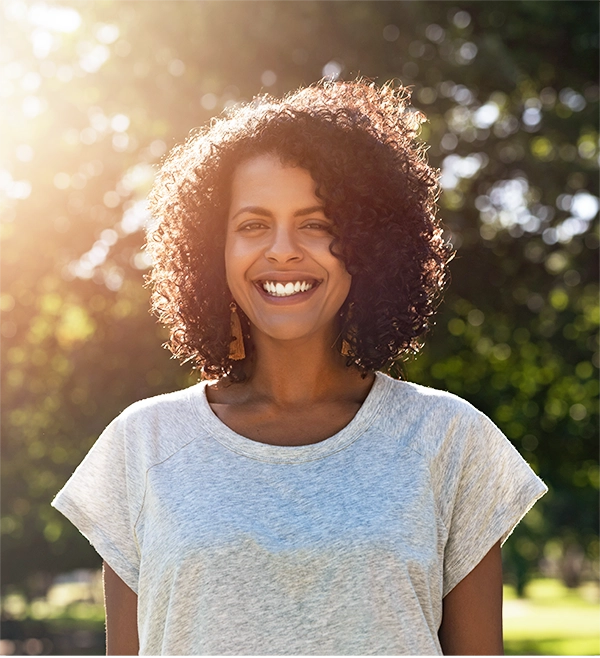 smiling woman with curly hair standing outside in bright, sunlit greenery, symbolizing long-term health and vitality.