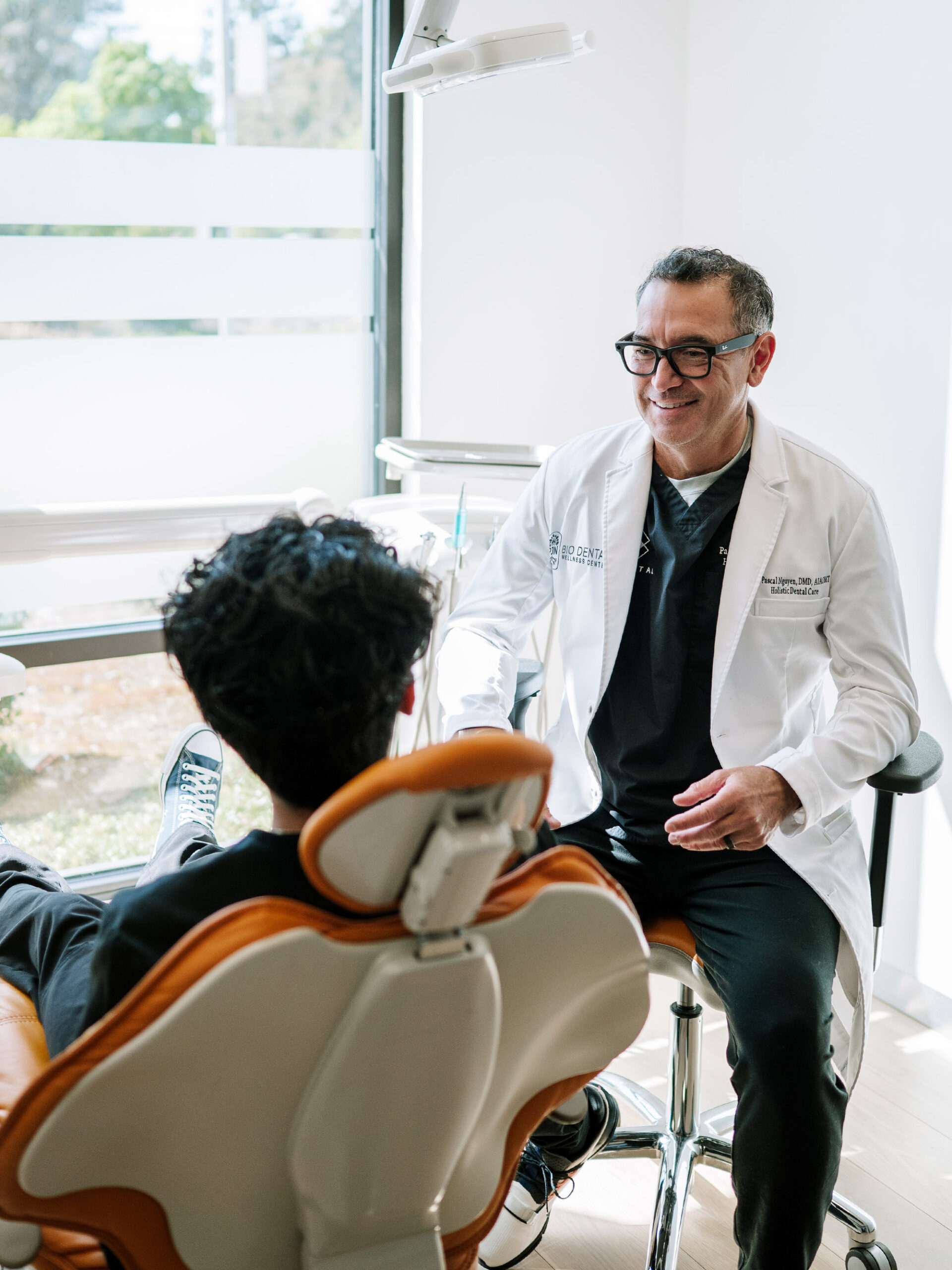 Dr. Pascal Nguyen sitting on a rolling stool, smiling and chatting face-to-face with a patient resting in an orange dental chair by a bright window.