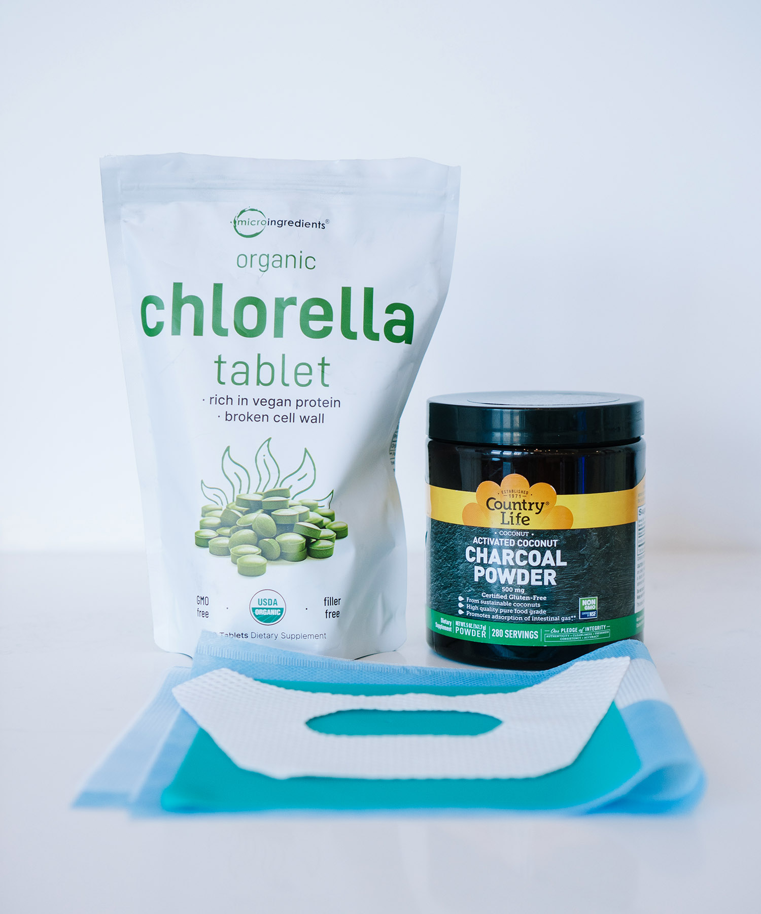 A bag of Micro Ingredients organic chlorella tablets, a jar of Country Life activated charcoal powder, and a protective blue dental drape used during a safe SMART mercury removal protocol.